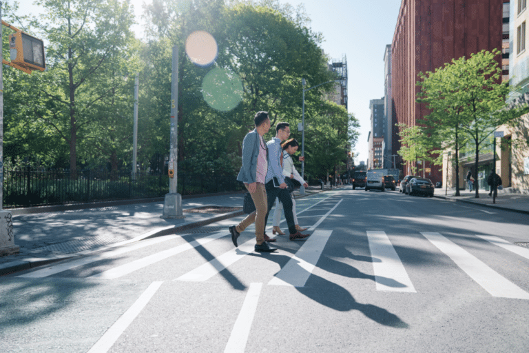 Three people walk across a crosswalk on a sunny day, with trees and buildings in the background.